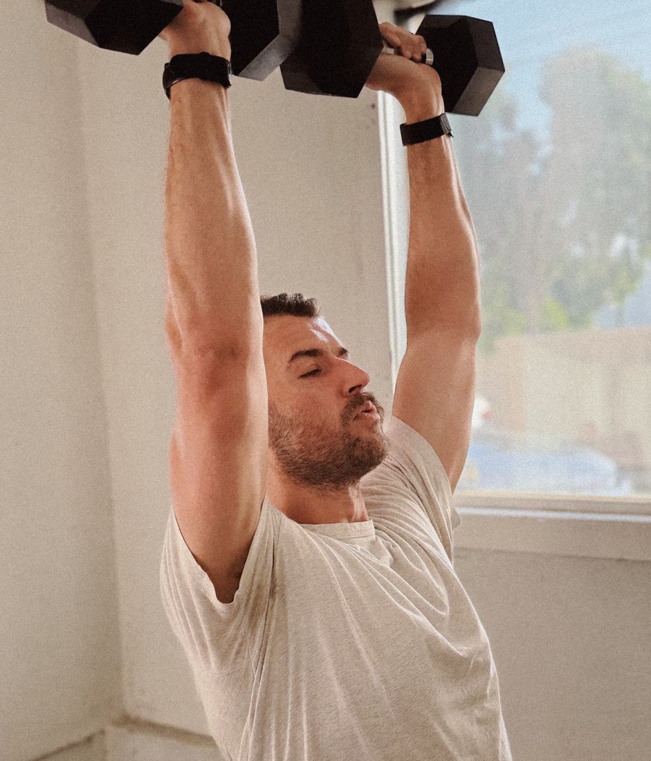 A man pressing dumbbells overhead beside a sunny window